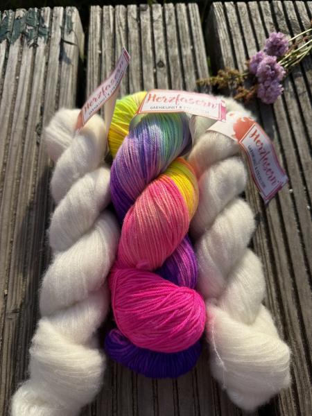 Handgefärbtes Garnset „Farbmagie“ von Herzfasern auf einer verwitterten Holzbank im Sonnenlicht fotografiert. In der Mitte liegt ein farbenfroher Strang Herzfasern No. 11 „Jolly“ mit Pink, Violett, Gelb und Türkis. Daneben liegen zwei fluffige Stränge Her